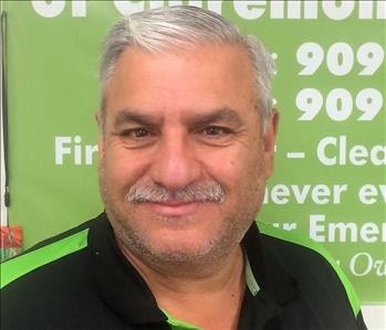 A seasoned gentleman with silver hair and mustache smiling in front of a SERVPRO banner. 