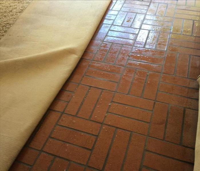 A beige carpet folded over, showing a red brick floor that is wet. 