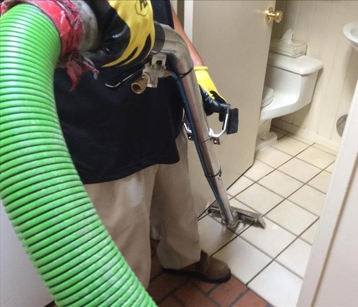 A water technician extracting water from a restroom with a silver wand and green hose in hand. 