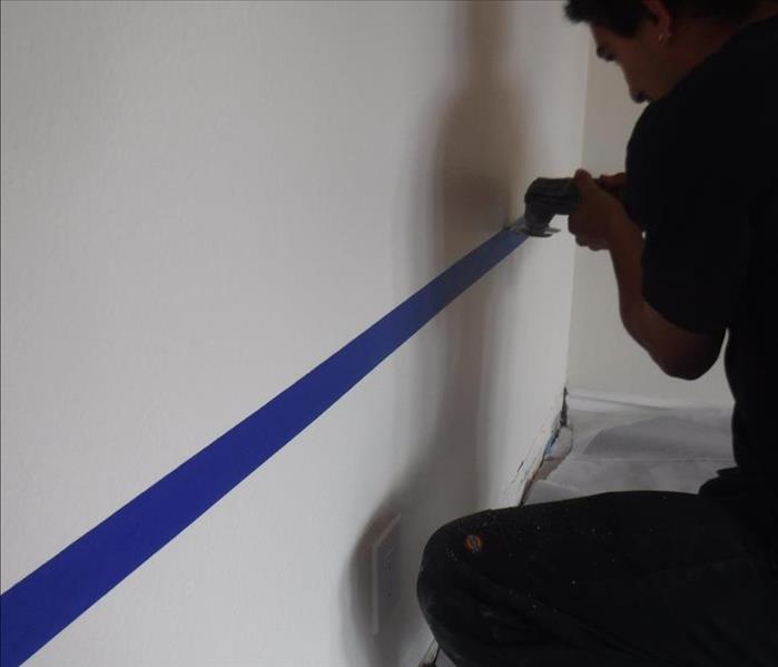 A SERVPRO of Claremont/Montclair technician carefully cutting drywall along a blue tape towards the bottom of the wall.